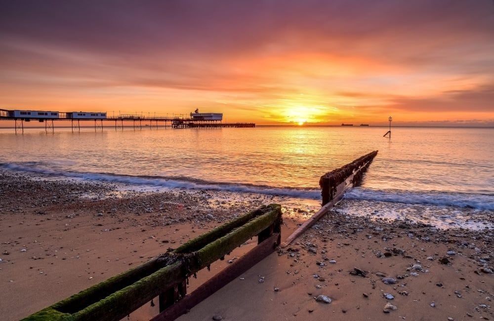 Sunrise over a beach with an old pier, calm sea, and pebbled shoreline.